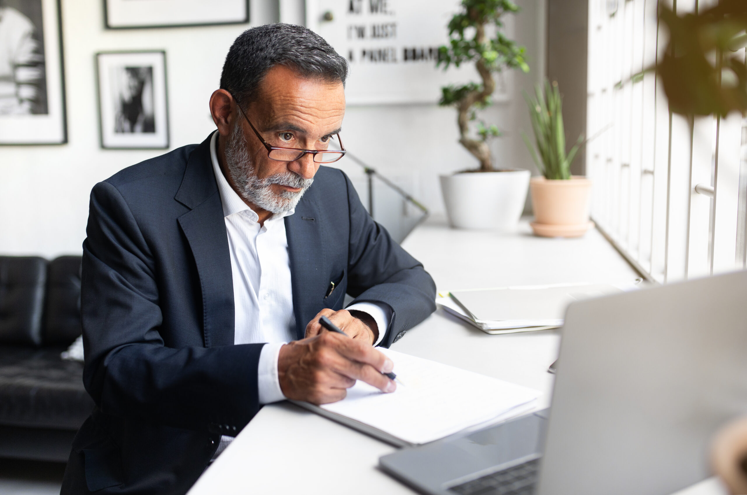 Busy serious caucasian senior businessman in suit makes notes in contract and looks at computer in modern office interior. Work remotely, business video call, meeting and webinar, study
